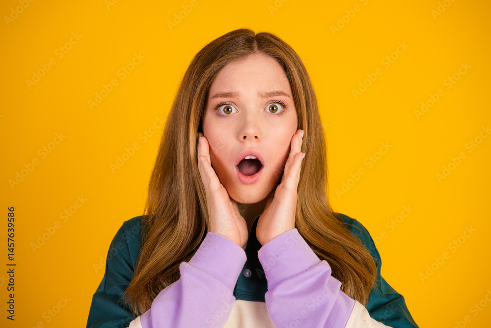 Surprised young woman posing against a vibrant yellow backdrop, expressing shock with her hands on her face
