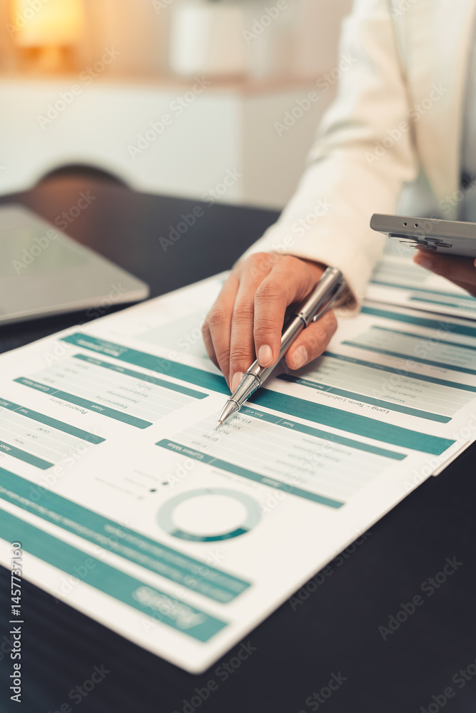Young female accountant hand holding business investment analysis pen. Accounting, finance expert working with financial data and calculations with graph paper document. Vertical close up.
