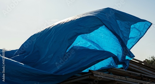 A large blue tarp billows dramatically in the wind, covering construction materials or a roof structure against a bright, sunny sky.