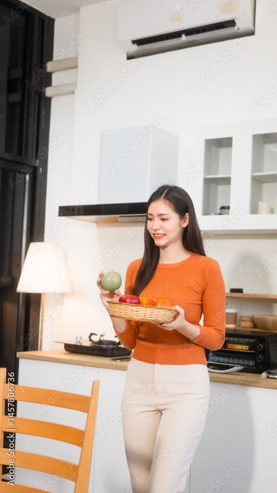 An Asian woman stands in the kitchen holding a basket of fresh fruits, ready to prepare a healthy meal. The kitchen is bright, and she radiates a sense of vitality