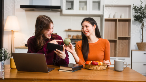 Two Asian women are sitting at a kitchen table, learning online at home. They smile happily while using laptops, surrounded by fruits and a coffee cup, enjoying their time