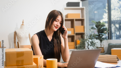 Happy Asian woman packing parcel boxes at a sofa table, smiling joyfully while checking online orders on laptop.She prepares products for delivery,managing her successful home-based online business