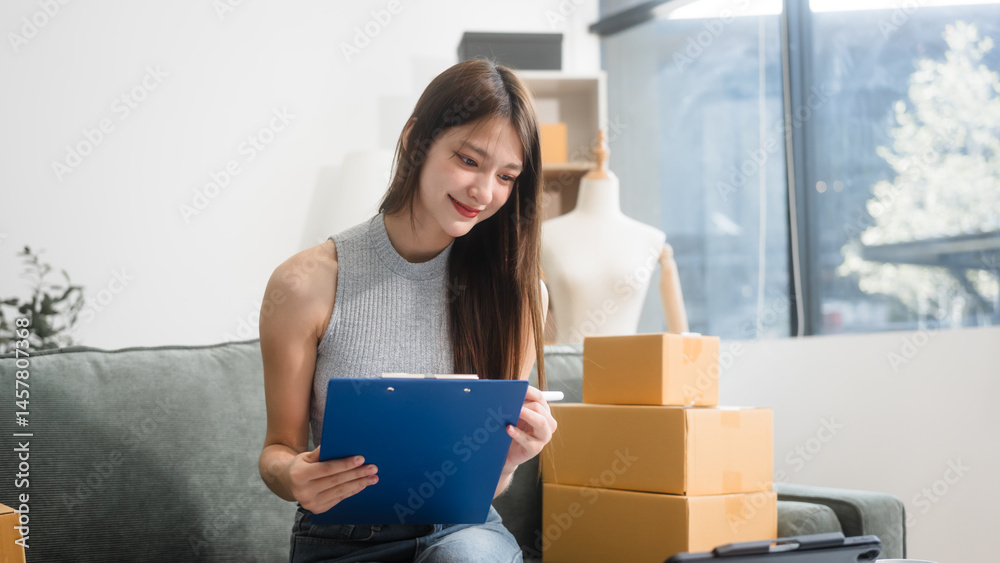 Happy Asian woman packing parcel boxes at a sofa table, smiling joyfully while checking online orders on laptop.She prepares products for delivery,managing her successful home-based online business