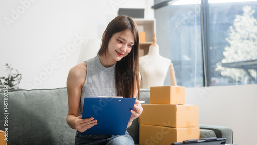 Obraz na płótnie Happy Asian woman packing parcel boxes at a sofa table, smiling joyfully while checking online orders on laptop.She prepares products for delivery,managing her successful home-based online business w salonie