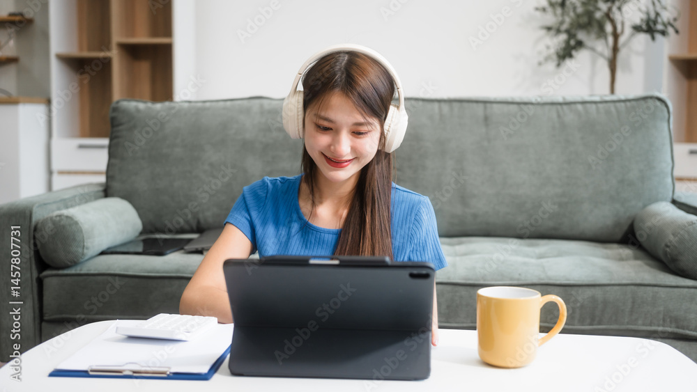 Asian woman sitting at a sofa table, wearing headphones and learning online at home.Focused on her laptop screen, she studies comfortably in a cozy environment,embracing digital education technology