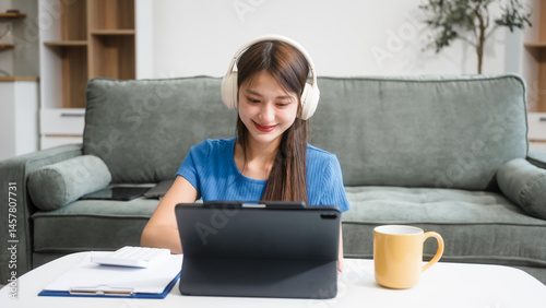 Obraz na płótnie Asian woman sitting at a sofa table, wearing headphones and learning online at home.Focused on her laptop screen, she studies comfortably in a cozy environment,embracing digital education technology w salonie
