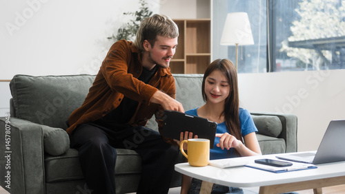 A Caucasian man and an Asian woman sit together at a table near the sofa, using a laptop at home. They appear relaxed and focused, working or browsing the internet together
