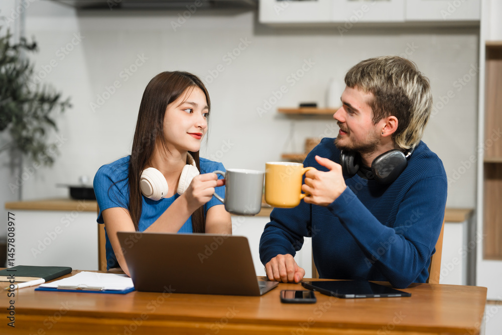Caucasian man and an Asian woman sit at a table at home, engaged in online learning. They share ideas, talk casually, and sip coffee, creating a relaxed and focused atmosphere