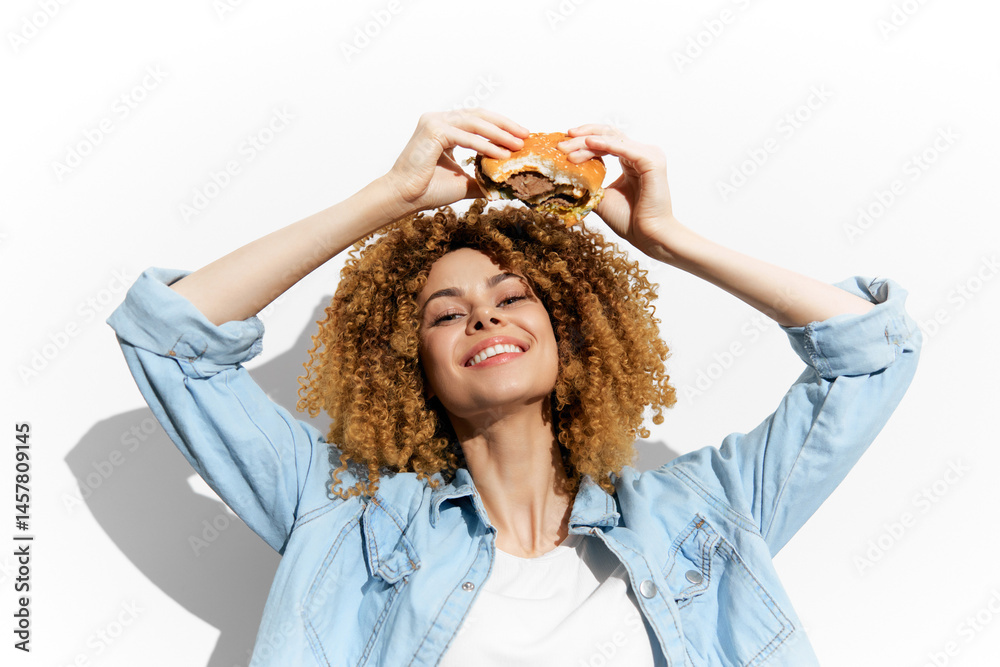 Happy woman with curly hair holding a burger, wearing a denim jacket against a white background, expressing joy and fun in a vibrant food themed concept