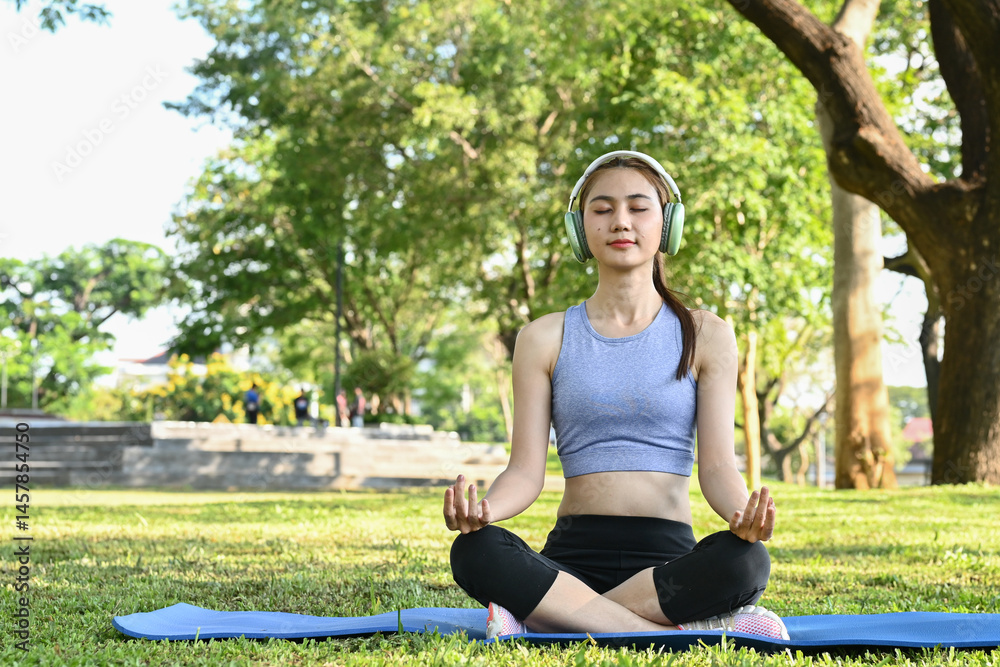 A woman is sitting on a mat in a park, wearing headphones and closed her eyes. She is in a relaxed and peaceful state of mind