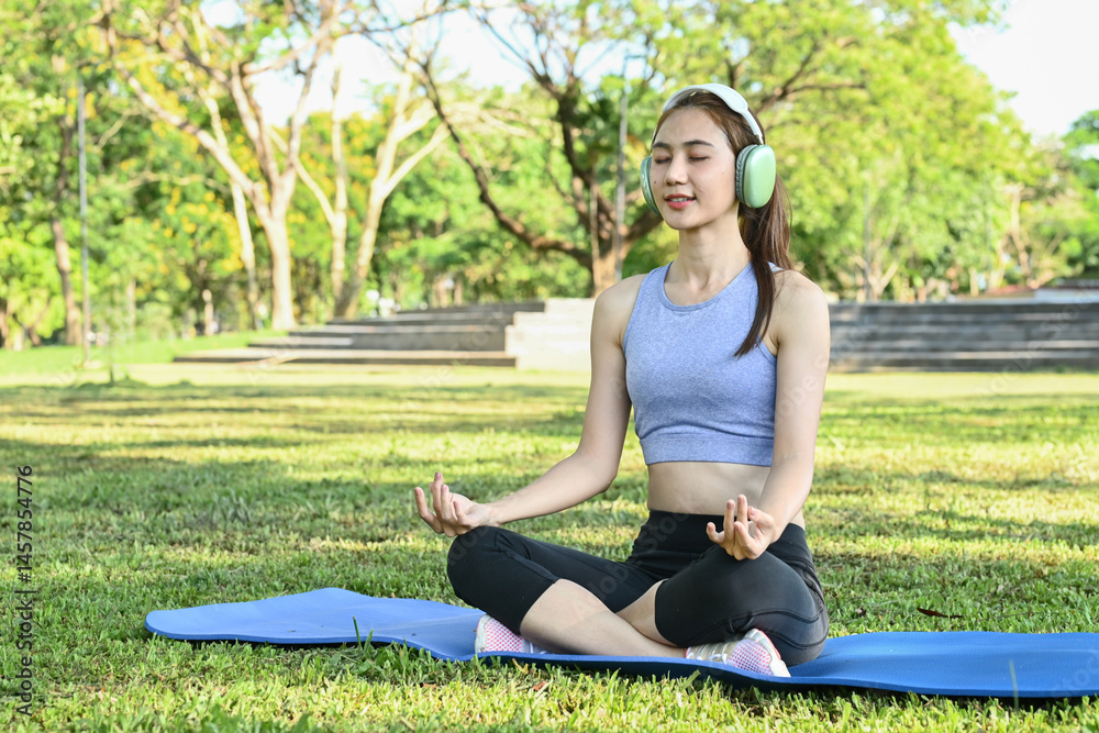 A woman is sitting on a mat in a park, wearing headphones and closed her eyes. She is in a relaxed and peaceful state of mind