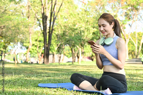 Obraz na płótnie A woman is sitting on a mat in a park, looking at her phone. She is wearing a blue tank top and black pants. w salonie