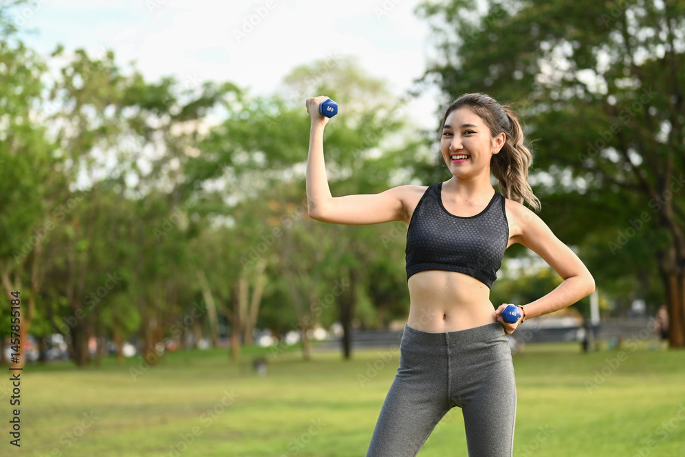 A woman is smiling and holding two dumbbells in a park. She is wearing a black tank top and gray pants.
