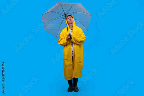 Man in rain boots and cloak posing with umbrella against vibrant blue backdrop, full length view.