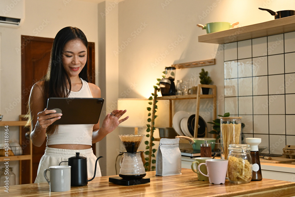 A woman is using a tablet in a kitchen. The kitchen is well-equipped with various items such as cups, bowls, and a bottle.