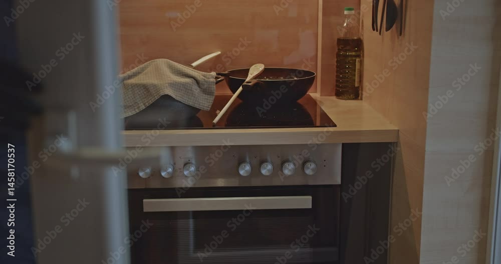 A man wearing an apron and gloves cooks and stirs food in a wok on the stove top. A second man opens and closes the refrigerator.
