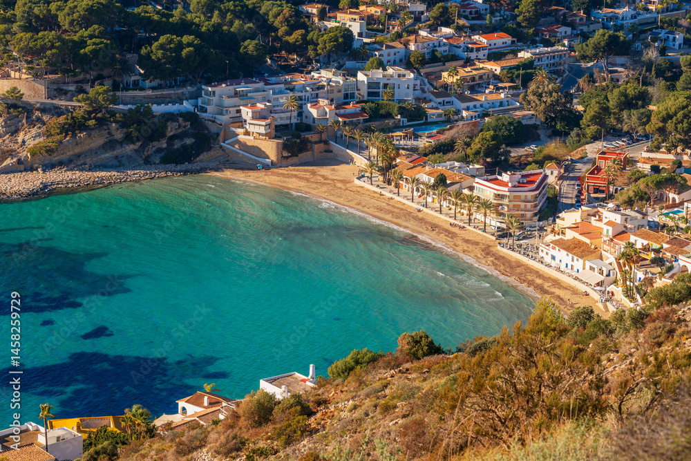 Fototapeta premium Clear turquoise water laps against the golden sand of El Portet beach in Moraira, Spain, as palm trees and Mediterranean houses line the peaceful bay in this elevated coastal view