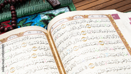 Bandung, Indonesia - 02.11.2025:  Photo of opened Holy Quran with Arabic calligraphy and wooden beads or tasbih with natural daylight and prayer mat below it on wooden background