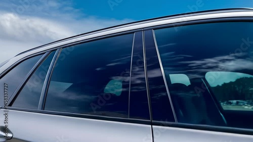 Modern silver SUV side profile with tinted windows against a bright blue partly cloudy sky showcasing clean lines and reflections of luxury