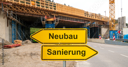 Zwei Wegweiser zeigen in unterschiedliche Richtungen mit dem Aufdruck Neubau und Sanierung vor einer Autobahnbrücke im Bau - FOTOMONTAGE