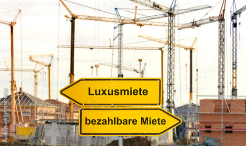 Vor einer Baustelle für ein Wohnhaus steht ein Wegweiser mit der Aufschrift Luxusmiete und bezahlbare Miete - FOTOMONTAGE