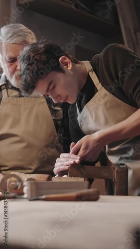 Caucasian man with gray beard guiding young male through woodwork. Both wearing aprons while focused on shaping piece of wood at workbench in cozy workshop filled with tools and materials.