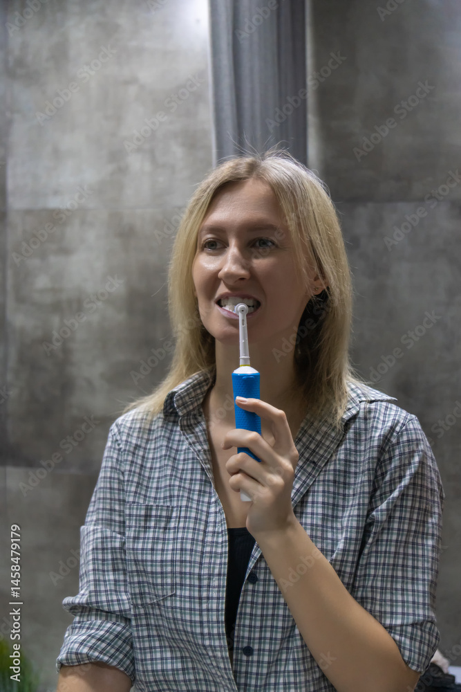 Young woman brushing her teeth with an electric toothbrush, maintaining good oral hygiene as part of her daily routine in a modern bathroom. Vertical photo