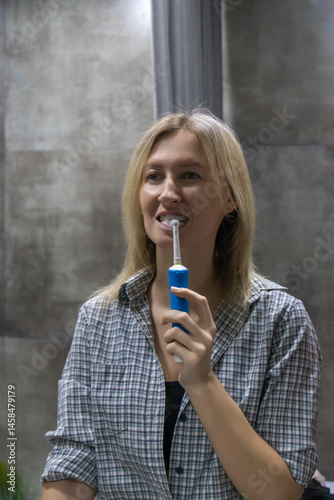 Obraz na płótnie Young woman brushing her teeth with an electric toothbrush, maintaining good oral hygiene as part of her daily routine in a modern bathroom. Vertical photo w salonie
