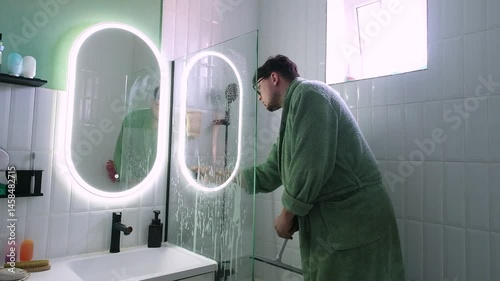 Man cleaning shower glass window in the bathroom with a scraper, using descaling spray