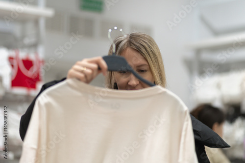 Smiling female customer enjoying a shopping experience in a fashion store, holding a hanger with a stylish white t-shirt while thoughtfully choosing from a variety of apparel