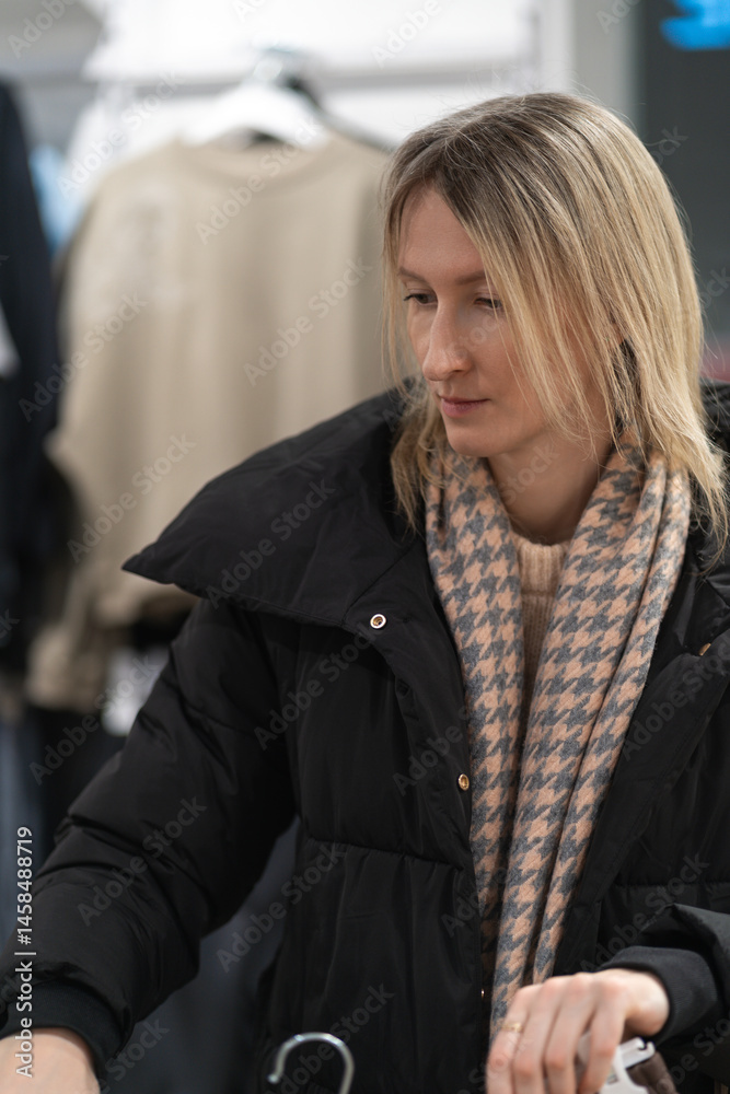 Young blonde woman dressed in stylish winter apparel, browsing and selecting garments from hangers in a chic fashion boutique, enjoying the shopping experience indoors. Vertical photo