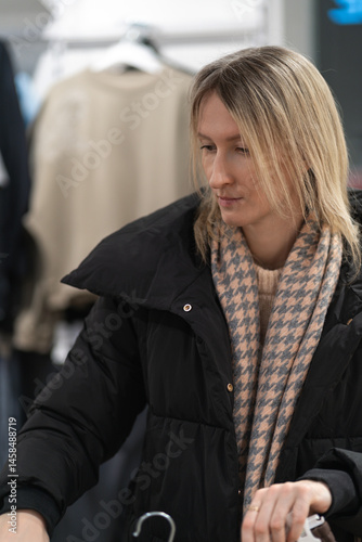 Young blonde woman dressed in stylish winter apparel, browsing and selecting garments from hangers in a chic fashion boutique, enjoying the shopping experience indoors. Vertical photo