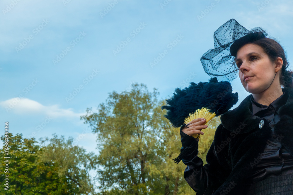 A confident and sophisticated lady in a strict black suit of the 19th century posts with a fan against the backdrop of an park.
