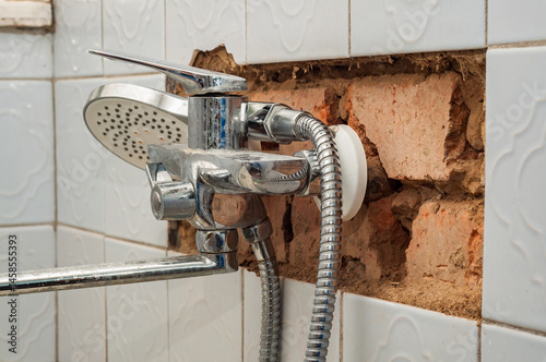 Chrome shower faucet protrudes from a broken tiled wall, revealing the brick structure behind.