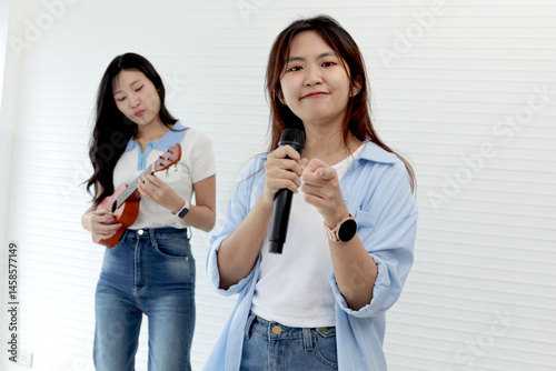 Two happy young Asian girls holding microphone and ukulele while they standing in living room. Cheerful teenage women enjoy singing and playing music together. People with music and instrument.