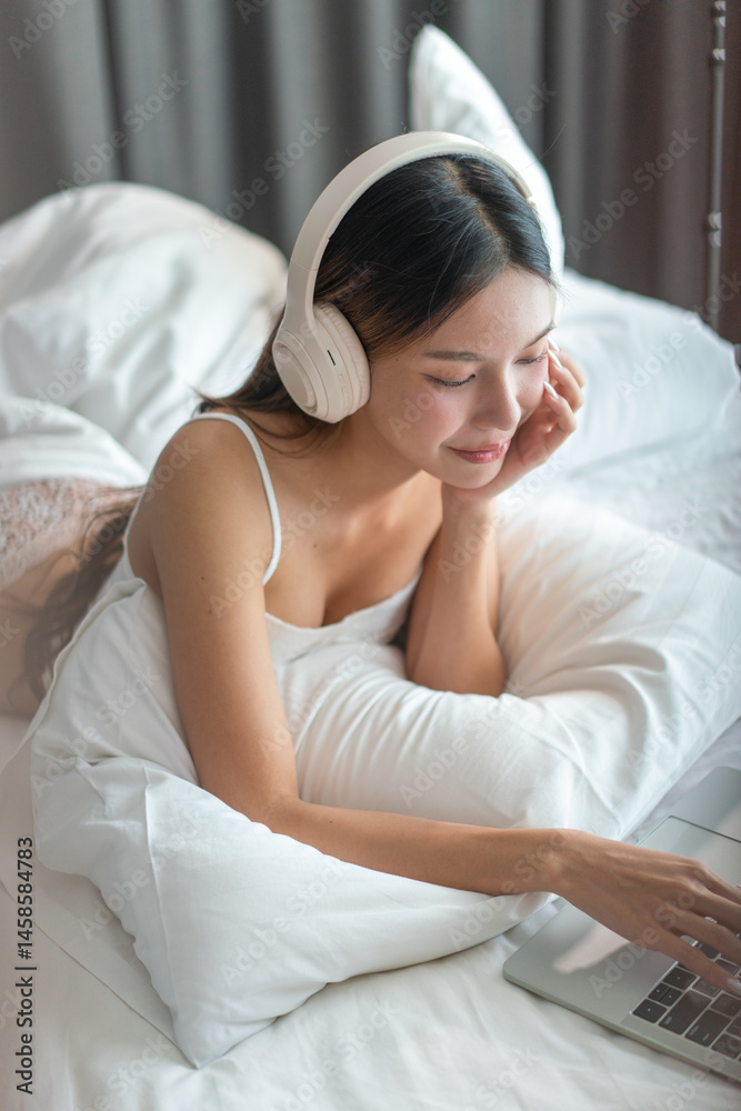 Asian woman relaxing on bed with headphones. Young female enjoying online content on laptop in cozy bedroom. Asian woman relaxing on bed with headphones. Young female enjoying online content on laptop in cozy bedroom.