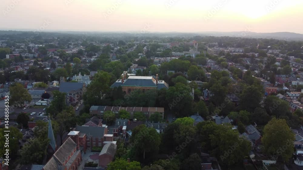 A wide drone shot over Lancaster, PA in summer, panning higher to reveal lush trees, city homes, glowing buildings, and a vibrant evening sky with distant mountains—capturing nature and urban charm.