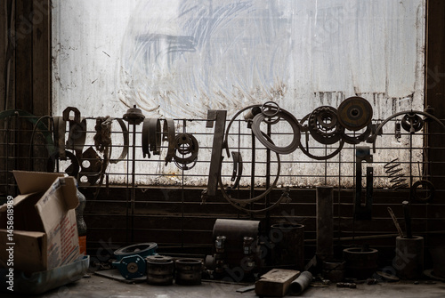 Obraz na płótnie Various greasy metal parts hang from a wire mesh against a dirty window, with more tools and components scattered on a workbench below. A cardboard box sits to the left. w salonie