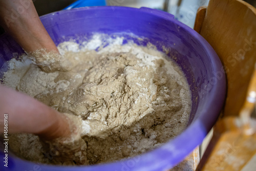 Hands kneading dough in a large plastic container during the process of preparing homemade bread. The dough is sticky and textured, showing active mixing.