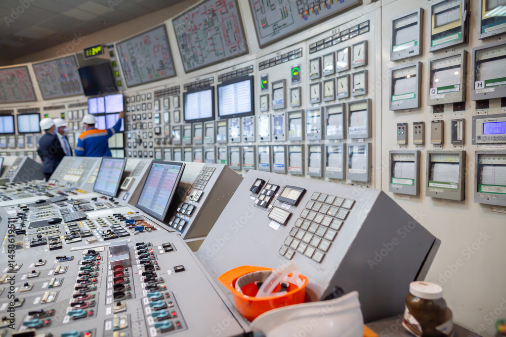 Control room with control panels featuring multiple dials, switches, and screens. Three workers in hard hats stand, one pointing at a wall-mounted screen, monitoring industrial operations.