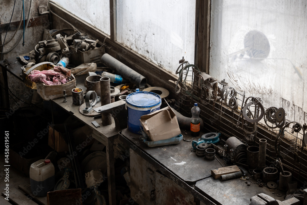 A cluttered workbench sits against a dirty window, covered with various metal parts, tools, containers, and discarded items in a messy workshop.