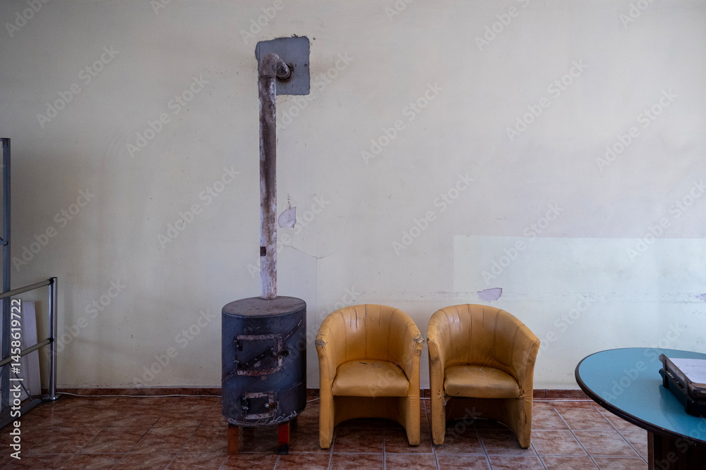 A simple waiting area features two worn yellow chairs beside a cylindrical metal stove with a chimney pipe against a pale yellow wall. A blue table is partially visible on the right.