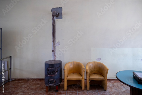 Obraz na płótnie A simple waiting area features two worn yellow chairs beside a cylindrical metal stove with a chimney pipe against a pale yellow wall. A blue table is partially visible on the right. w salonie