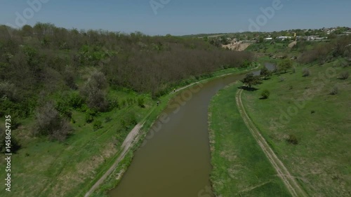 Tracking drone view of cyclists racing on a dirt road along a river and near young forest with green grass