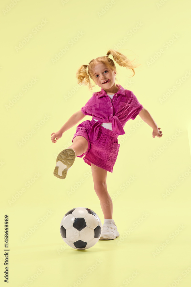 Smiling girl in pink outfit joyfully playing with soccer ball on pastel yellow background. Active lifestyle of kid. Concept of childhood, summer activity, sports joy and leisure