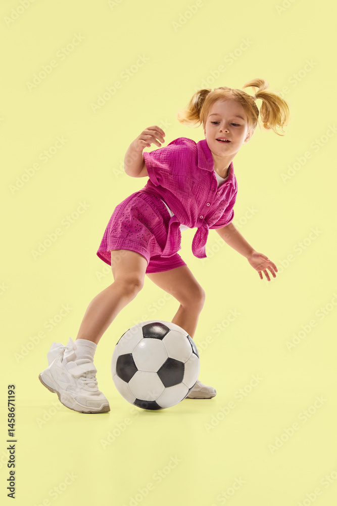 Joyful girl, child controls soccer ball with foot while playing against pastel yellow studio background. Game on. Concept of childhood, summer vacation, active lifestyle, fun, sport