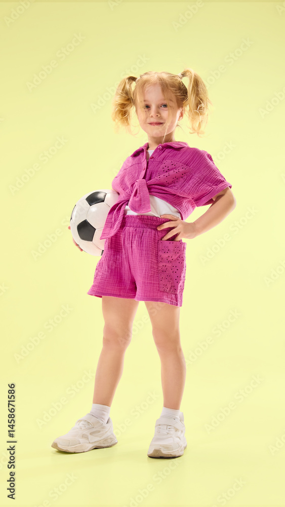 Smiling child girl posing in pink shorts and shirt with soccer ball on hip against light yellow background. Concept of childhood, summer vacation, active lifestyle, fun, sport