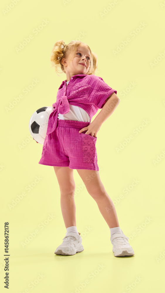 Full-length low angle view of happy smiling child girl wearing pink summer outfit, posing with soccer ball against yellow background. Concept of childhood, summer vacation, active lifestyle, sport