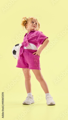 Obraz na płótnie Full-length low angle view of happy smiling child girl wearing pink summer outfit, posing with soccer ball against yellow background. Concept of childhood, summer vacation, active lifestyle, sport w salonie