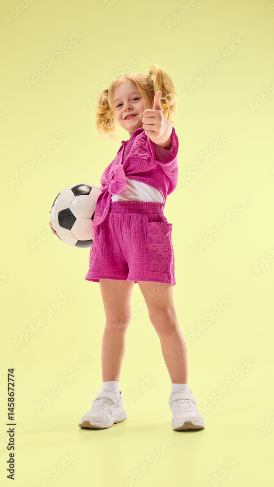 Low angle view of happy child girl in pink shorts and shirt, standing with soccer ball and giving thumbs up against yellow background. Concept of childhood, summer vacation, active lifestyle, sport